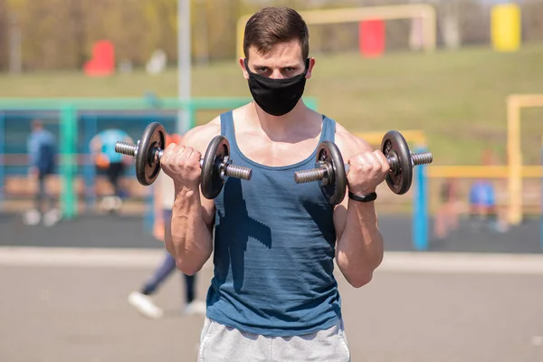 Young sports guy doing exercises with dumbbells in a medical mask on the playground during a pandemic. COVID-19. Health care.