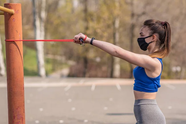 A young and athletic girl in a medical mask does exercises with sports elastic bands during a pandemic. COVID-19. Health care.