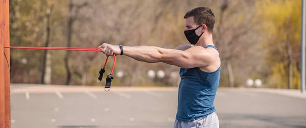 A young and athletic guy in a medical mask does exercises with sports elastic during a pandemic. COVID-19. Health care.