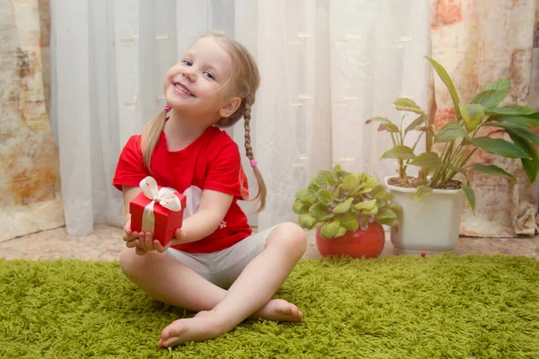 En vacker blond liten flicka sitter på en matta hemma och håller en gåva i händerna. Upphovsrätt — Stockfoto