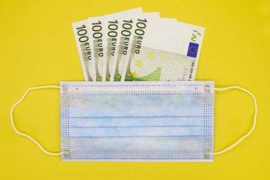 Tıbbi korumalı mavi maske, sarı arka planda 100 Euro 'luk banknotların üzerinde yatıyor. Seçici odaklanma. Yüz maskeleri için fiyat artışı. Mesaj için yer var. Kapat..