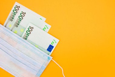 Tıbbi koruyucu cerrahi mavi maske, turuncu arka planda 100 Euro 'luk banknotların üzerinde yatıyor. Seçici odaklanma. Yüz maskeleri için fiyat artışı. Mesaj için yer var. Kapat..