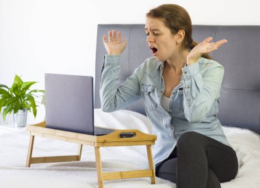 Güzel, genç, şaşırmış ve korkmuş bir kadın. Ağzı ve kolları açık bir şekilde evde, bilgisayarlı, yaşam tarzlı, fotokopi çekilen bir yatakta tele çalışıyor. Coronavirus konsepti sırasında serbest ve merkez ofis