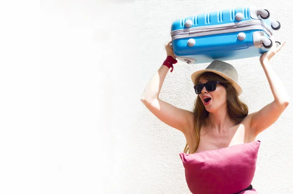 Excited screeming woman in red pillow dress, glasses and summer hat with blue luggage on her head on white wall background. Vacation time. Pillow Challenge due to stay home isolation. Caronavirus Quarantine.