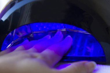 Ultraviyole lamba içindeki bir kadının eli yakın plan çekilmiş. Evde yeni sürdüğü jel ojesini iyileştiriyor. Ev yapımı tırnak bakımı konsepti