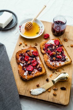 Ballı kızarmış ekmek, krem peynir ve kırmızı meyve reçeli, masa ve ahşap üzerinde.