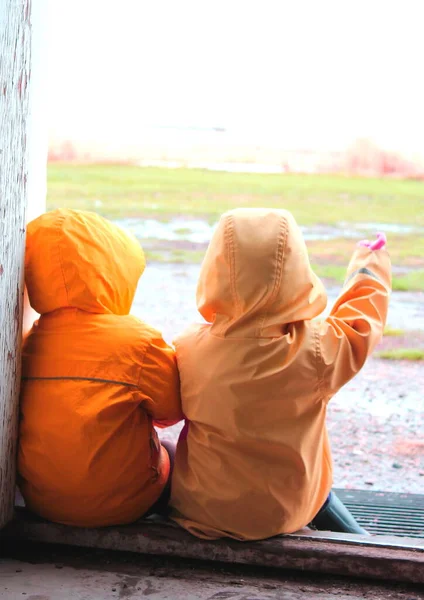 Rain jackets being worn as two young children playing in the rain wait on a door step for the spring weather to pass on a country setting.