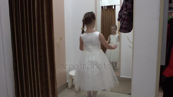 5 jaar oud meisje proberen op een witte jurk in een kledingwinkel. — Stockvideo