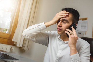 İş adamı, bir sorun yüzünden elini alnının yanına koyarken iş yerindeki bilgisayarının önünde telefonla konuşuyor. Bir şeyi unutuyor ya da kendini kötü hissediyor. Stres ve tükenmiş iş kavramı.