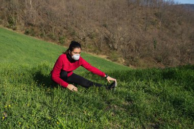 Genç sportif kadın, Coronavirus için yüz koruyucu maskeyle koşuyor ve esneme egzersizleri yapıyor. Spor açık hava ve salgın konsepti.