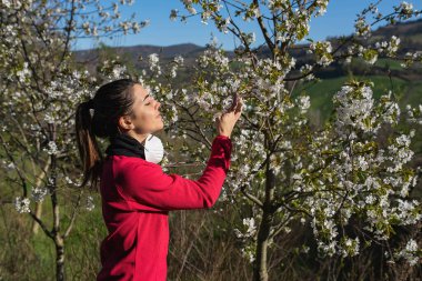 Koronavirüsün çiçek koklamasını önlemek için koruyucu maskesini çıkaran sportif bir kadın. Salgın ve doğa kavramı.