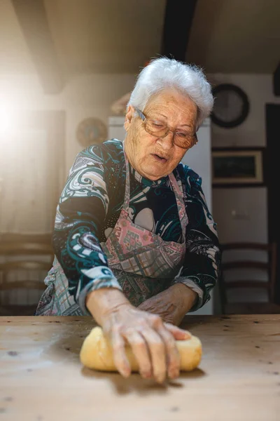 Caucasian grandmother, senior old woman cooking in her kitchen traditional recipes of Italian cuisine. Fresh handmade pasta with eggs and flour, rolling dough on wooden cutting board.