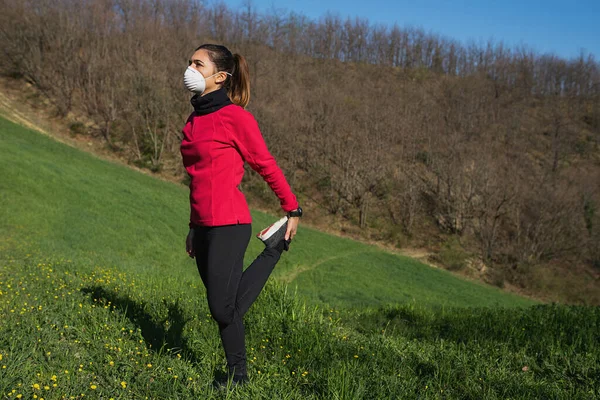 Genç sportif kadın, Coronavirus için yüz koruyucu maskeyle koşuyor ve esneme egzersizleri yapıyor. Spor açık hava ve salgın konsepti.