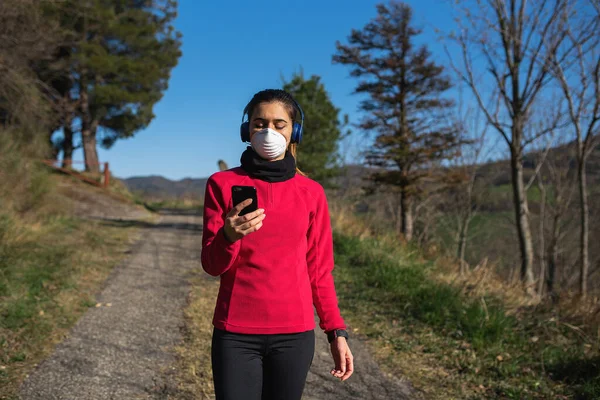 Kulaklıklı ve akıllı telefonlu sportif bir kadın açık havada Coronavirus sırasında yüz koruyucu maskeyle spor yapıyor. Salgın, sağlıklı yaşam tarzı, spor anlayışı.