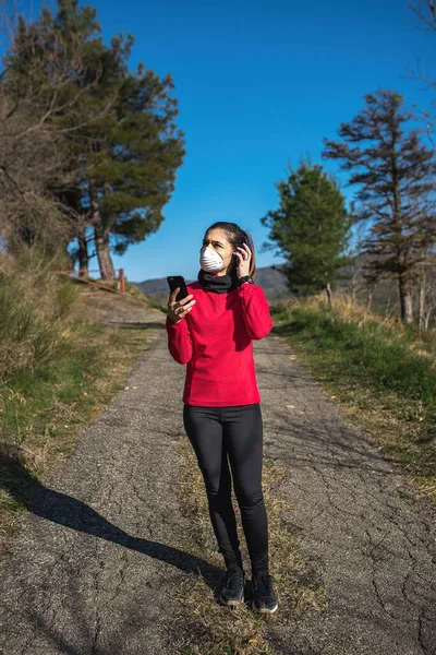 Kulaklıklı ve akıllı telefonlu sportif bir kadının koronavirüs sırasında yüz koruma maskesiyle açık havada spor yaptığı dikey görüntü. Salgın, sağlıklı yaşam tarzı, spor anlayışı.