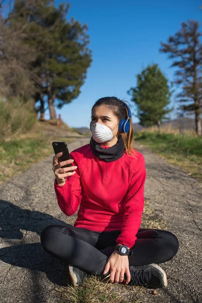 Sportive woman sitting on the ground outdoor after fitness and jogging, listening to music with headphones and mobile wearing protective face mask during coronavirus pandemic.