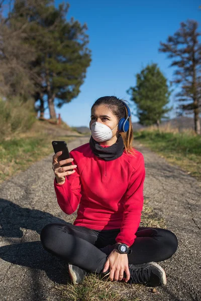 Spor kadın spor salonunda spor yapıyor, spor yapıyor, kulaklıkla müzik dinliyor ve koronavirüs salgını sırasında koruyucu yüz maskesi takıyor..