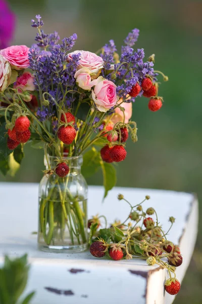 Juicy fresh bouquet of strawberries, lavender and roses stands on a white chair in a jar of water. Delicious berries and aromatic herbs collected in the garden and in the forest. Vertical frame