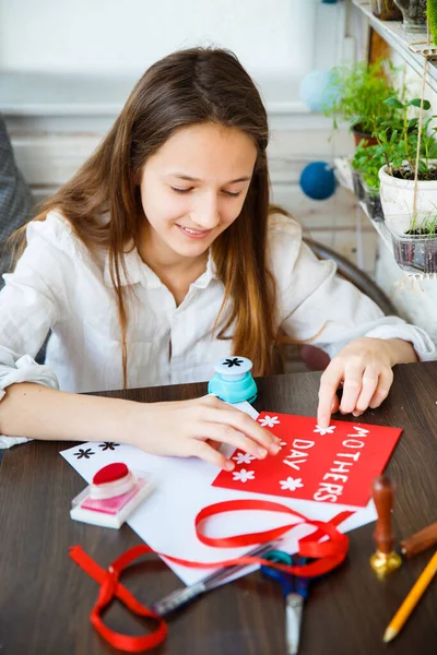 A teenage girl makes a handmade card for mother's day holiday. On a wooden table are stationery - scissors, tape, pencil, paper, a hole punch for flowers. Vertical frame