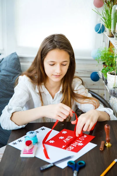 A teenage girl makes a handmade card for mother\'s day holiday. On a wooden table are stationery - scissors, tape, pencil, paper, a hole punch for flowers. Knots a red ribbon. Vertical frame