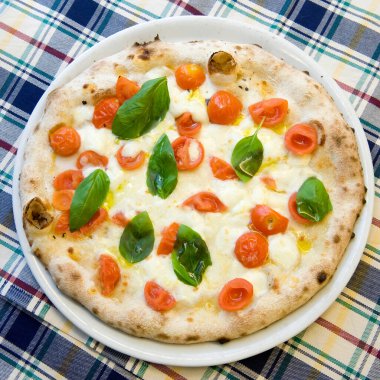 Gerçek bir İtalyan sebze pizzası, kiraz domatesli, mozzarella ve fesleğenli, ekose masa örtüsünün üstünden manzaralı.