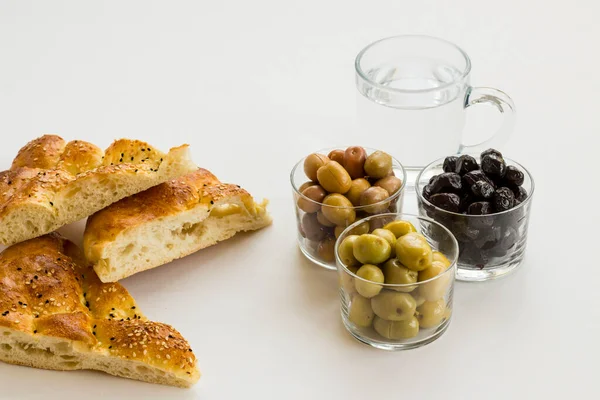 Beyaz zemin üzerinde taze Ramazan ekmekleri, zeytin çeşitleri ve bir bardak su ile dilimlenmiş..