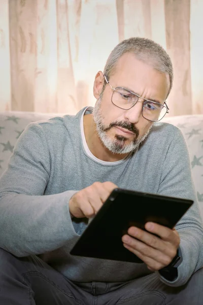 Oturma odasından çalışırken elinde tableti tutan gözlüklü bir adam.