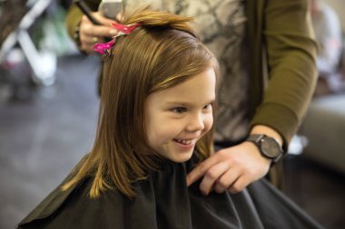 Küçük kız kuaförde oturuyor, siyah pelerinli.