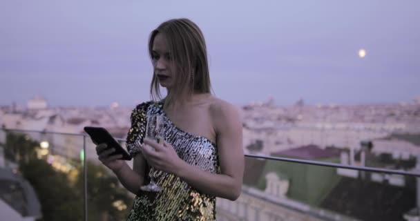 Кавказская женщина в коктейльном платье на крыше пьет шампанское и проверяет смартфон — стоковое видео