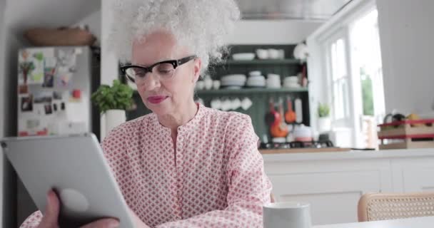 Mulher idosa adulta olhando para tablet digital enquanto toma café da manhã — Vídeo de Stock