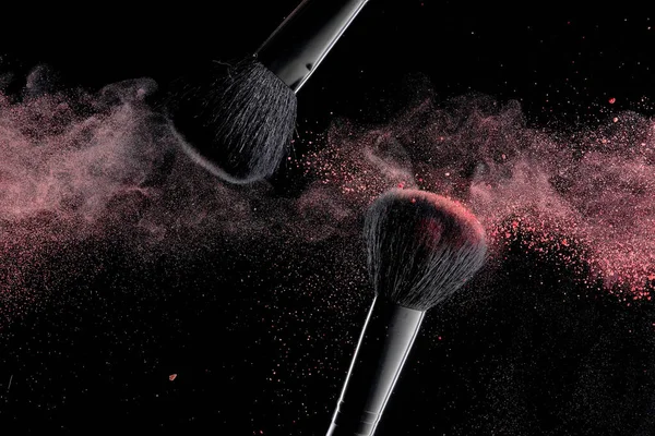 Makeup brushes with colorful powder explosion on black background. High speed photography of cosmetics.