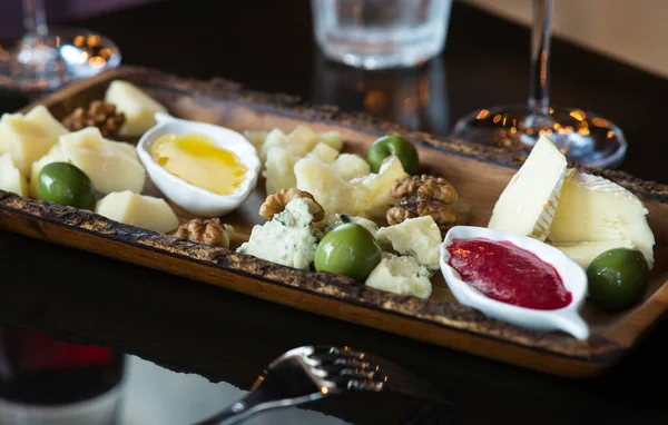 Yeşil zeytin, fındık, bal ve reçelli peynir tabağı. İki şarap bardağı, metal çatal. Siyah parlak masa, ahşap tabak.