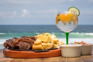 Picanha, Brezilya 'nın geleneksel et dilimi, sıcak bir tabakta kızarmış kasava ile servis edilir. Arka planda tropik bir içecek ve plaj var..