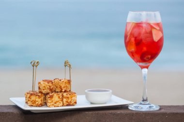 Rennet peyniri ve muhallebi unundan yapılan ve biber jölesiyle servis edilen Dadinho de tapioca adlı atıştırmalık. Arka planda tropik içki ve plaj var..