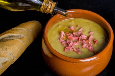 Bezelye çorbasının içine zeytinyağı dökülüyor. Siyah arka planda domuz pastırması ve ekmek ekmeği var..