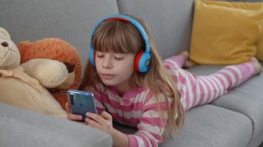 Kulaklıklı şirin küçük bir kız kanepede akıllı bir telefonla oynuyor.