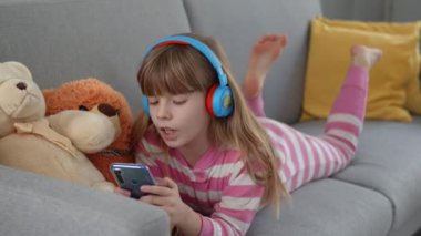 Kulaklıklı şirin küçük bir kız kanepede akıllı bir telefonla oynuyor.