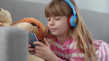 Kulaklıklı şirin küçük bir kız kanepede akıllı bir telefonla oynuyor.