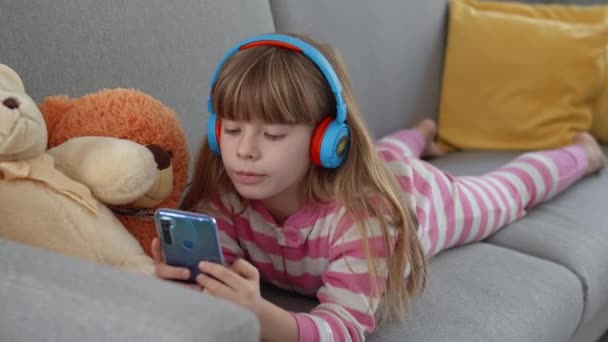 可爱的小女孩带着耳机在沙发上的智能手机上玩耍 — 图库视频影像