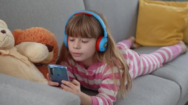 Linda Niña Con Auriculares Juega Teléfono Inteligente Sofá — Vídeo de stock