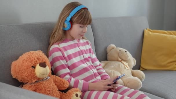Cute Little Girl Headphones Plays Smartphone Sofa — Stock Video