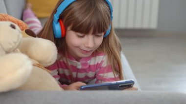 Kulaklıklı şirin küçük bir kız kanepede akıllı bir telefonla oynuyor.