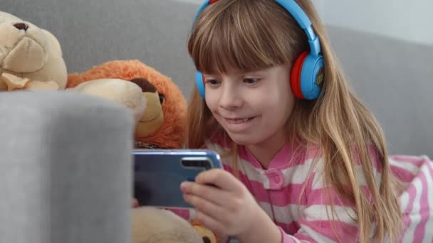 Kulaklıklı Şirin Küçük Bir Kız Kanepede Akıllı Bir Telefonla Oynuyor — Stok video