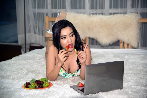 Portrait of sexy young Asian woman lying on bed and working at laptop. — Stock Photo, Image