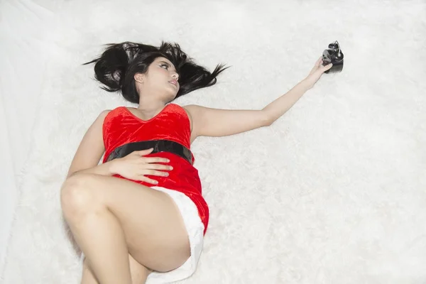 Imagen retrato de una hermosa mujer asiática en Santa Claus roja posando tumbada con reloj en la cama en una habitación . — Foto de Stock