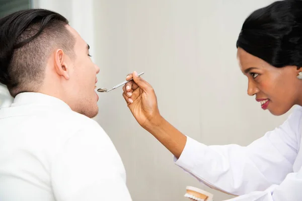 Dentista Africano feminino verificando dentes de paciente do sexo masculino olhando no espelho dental. Conceito de procedimento oral . — Fotografia de Stock