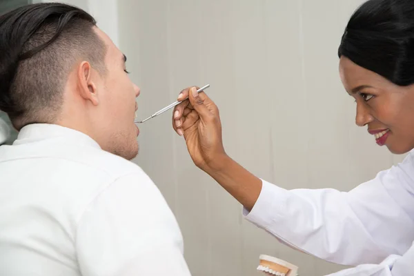 Dentista Africano feminino verificando dentes de paciente do sexo masculino olhando no espelho dental. Conceito de procedimento oral . — Fotografia de Stock
