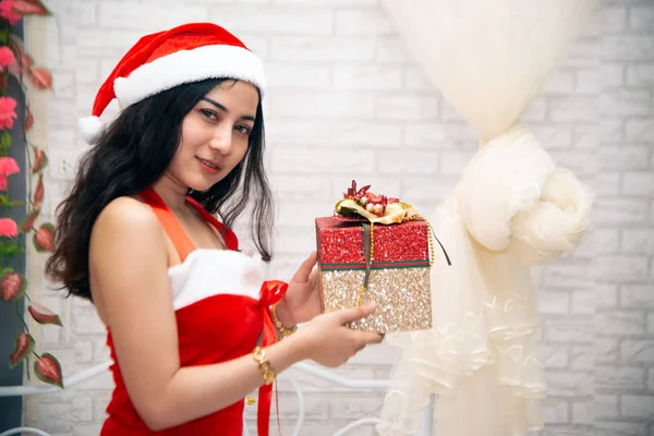 Una hermosa mujer asiática con un vestido rojo y un sombrero de Santa Claus está sentada y sonriendo, sosteniendo una caja de regalo de Navidad. Realizar diferentes posturas . — Foto de Stock
