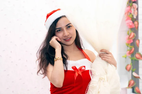 Piękna Azjatka ubrana na czerwono i nosząca czapkę Mikołaja stoi i uśmiecha się na Boże Narodzenie. I działać na różne sposoby.. — Zdjęcie stockowe