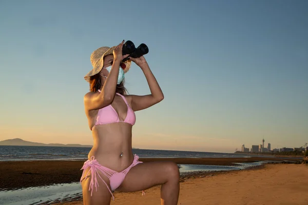 A senior Asian woman wearing a pink bathing suit, wearing glasses and wearing a straw hat, with a nose mask, anti-virus COVID-19, and anti-PM2.5 dust posing happily holding Binoculars on the beach in evening on a blue sky.